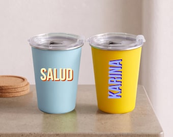 Puede incluir: Dos vasos de acero inoxidable con tapas de plástico transparente. El vaso izquierdo es azul claro con la palabra SALUD impresa en naranja y amarillo. El vaso derecho es amarillo con el nombre KARINA impreso verticalmente en azul y lila.