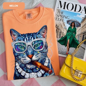 Op de afbeelding: Een meloenkleurig t-shirt met een afbeelding van een blauwe kat die een met diamanten ingelegde bril en een bijpassende ketting draagt terwijl deze een sigaar rookt. Het t-shirt heeft een Comfort Colors label in de nek en is maat L.