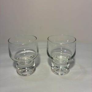 May include: Two clear glass tumblers with a thick, rounded base and a slightly wider, cylindrical upper section. The glasses feature a smooth, transparent finish with a heavy bottom design, providing a stable structure for these drinking vessels.