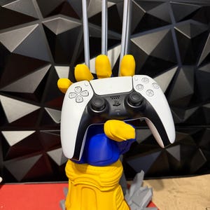 Pode incluir: Um suporte de comando em forma de mão amarela e azul com três longas garras prateadas que se estendem dos nós dos dedos. O suporte segura um comando de jogo sem fios branco e preto com um logótipo central PlayStation e botões direcionais.