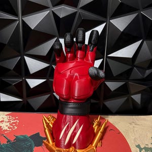May include: A red, upright hand sculpture featuring black fingertips and a black wrist band. The base of the hand is decorated with gold-colored bullet casings and three prominent white claw marks etched into the red surface of the wrist area.