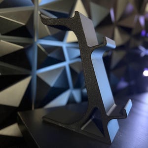 May include: A black, textured, vertical stand with a curved top support and a lower shelf, featuring a triangular cutout at the base. The stand has a matte finish and is designed with multiple tiers to hold items securely in an upright position.