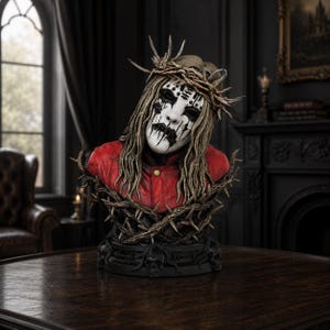 Puede incluir: Un busto que representa una figura con una máscara blanca con marcas negras, cabello largo y una corona de espinas. La figura viste una chaqueta roja y se asienta sobre una base decorada con calaveras negras y ramas espinosas.