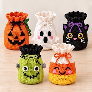 May include: Five crocheted drawstring pouches featuring Halloween designs: an orange pumpkin, a white ghost, a black cat, a green monster with stitches, and a yellow, orange, and white candy corn, all with cinched tops and decorative ties.