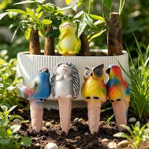 Puede incluir: Un conjunto de cuatro estacas decorativas de jardín con un pájaro azul, un erizo gris, un búho amarillo y un loro multicolor, cada uno con una base cónica. Una figura de rana verde se encuentra al fondo dentro de una maceta blanca estriada.