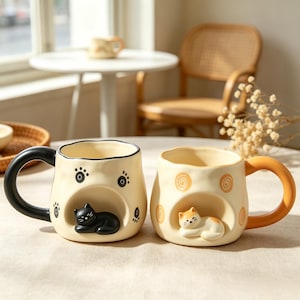 Puede incluir: Dos tazas de cerámica de color crema con bordes ondulados e irregulares. La taza de la izquierda tiene un asa negra, patrones de huellas negras y un gato negro esculpido descansando en un hueco. La taza de la derecha tiene un asa naranja, patrones de espirales naranjas y un gato blanco y naranja esculpido descansando en un hueco.