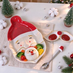 Puede incluir: Un plato de cerámica con forma de Santa Claus con gorro rojo y barba blanca, acompañado de un cuenco para condimentos con forma de bigote y una cuchara de metal que presenta un pequeño gorro de Santa en el extremo.