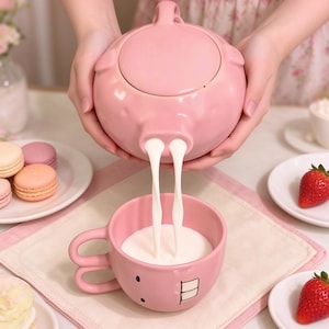 Puede incluir: Una tetera de cerámica rosa con forma de cerdo vierte un líquido blanco en una taza de cerámica a juego. La taza presenta una cara de dibujos animados con dos dientes frontales prominentes y un asa en bucle, mientras que la tetera tiene un pico con forma de hocico.