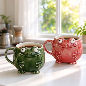 Può includere: Due tazze in ceramica a forma di rana, una in verde bosco scuro e l'altra in un vivace rosa rossastro. Ogni tazza presenta un corpo arrotondato, un manico curvo e un volto di rana scolpito con occhi prominenti e un ampio sorriso.