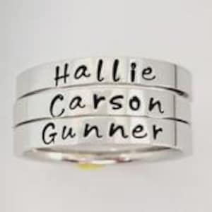 May include: A stack of three polished silver-toned metal rings. Each ring features a smooth, flat band with the name Hallie, Carson, or Gunner engraved in a black, cursive-style font on the front surface of the metal band.
