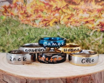 May include: A collection of six metal rings featuring engraved names. The rings include a silver-tone ring with Zack, a silver-tone ring with Easton, a blue ring with James, a gold-tone ring with Hosea, a camouflage ring with Tristan, and a silver-tone ring with Cole.