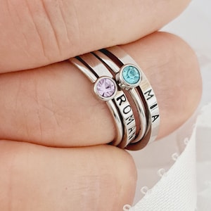 Puede incluir: Tres anillos de metal plateado apilados. Cada anillo presenta un pequeño cristal redondo engastado en azul claro, rosa claro y transparente. Dos de los anillos están grabados con los nombres ROMAN y MIA en letras mayúsculas negras.