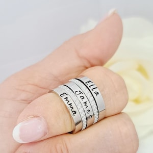 May include: Three stacked silver-toned rings featuring clear stones along the edges. Each ring is engraved with a name in black script: Emma, James, and Ella. The rings have a smooth, polished finish and are worn together on a single finger.