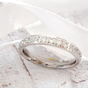 May include: A silver-toned metal band ring featuring a row of small, clear, round-cut stones set along the top half of the band. The ring has a polished finish and is positioned in front of a white ribbon, showcasing a simple and clean design.