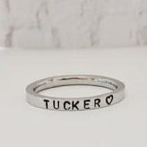 May include: A thin, polished silver-toned metal band ring featuring the engraved black text TUCKER followed by a small heart symbol. The ring has a flat, smooth surface and a simple, circular design, showcasing clear, legible lettering on the outer band.