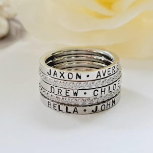 以下が含まれることがあります： シルバー調のリング5本のスタック。3本は滑らかなバンドで、JAXON、AVERIE、DREW、CHLOE、BELLA、JOHNという名前が刻印されています。2本は小さな透明で輝く石がちりばめられており、刻印されたバンドと交互に重なっています。