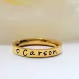 Puede incluir: Un anillo de banda en tono dorado con un acabado liso y pulido. La superficie exterior del anillo está grabada con un pequeño símbolo de corazón seguido del nombre Carson en una fuente de escritura simple, oscura y en minúsculas, centrada.