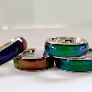 Könnte beinhalten: Vier Stimmungsringe mit metallisch silbernen Bändern und farbwechselnden Zentren. Die Ringe zeigen verschiedene Farbtöne, darunter tiefes Violett, dunkles Orange und leuchtendes Grün, mit einer glatten, polierten Oberfläche.