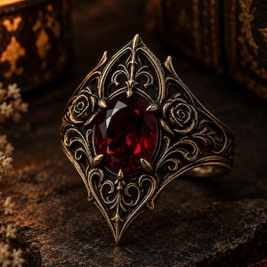 May include: A ring featuring an oval-cut deep red gemstone set in an antiqued gold-tone metal band. The metalwork displays intricate filigree patterns, pointed architectural details, and sculpted rose motifs on both sides of the central stone.