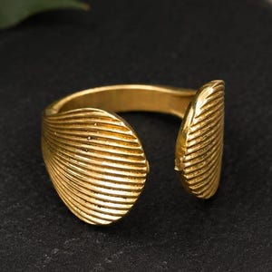 May include: A gold-toned open ring featuring two shell-shaped ends with a textured, ribbed surface. The band is smooth and circular, designed with a gap between the two decorative shell elements, creating a distinct, symmetrical appearance.