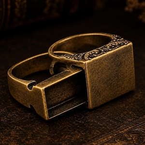 Puede incluir: Un anillo de metal en tono latón antiguo que presenta un compartimento deslizante rectangular y hueco. La superficie superior del anillo está decorada con intrincados patrones de volutas en relieve a lo largo de los bordes, dando un acabado texturizado.