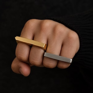 May include: Two horizontal bar rings worn on adjacent fingers. One ring features a polished gold-tone finish, while the other ring has a brushed silver-tone finish. Both rings have a flat, rectangular top surface and a smooth, solid metal band design.
