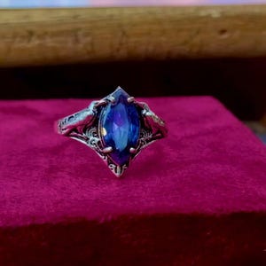 May include: A silver-toned metal ring featuring a marquise-cut, deep blue gemstone set in the center. The band displays intricate, raised metalwork patterns that extend from the setting along the sides of the ring's shank.