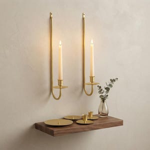 May include: Two gold-toned metal wall-mounted candle holders with long, slender arms, each holding a cream-colored taper candle. Below, a wooden shelf displays three small gold-toned candle plates and a glass vase with eucalyptus stems.