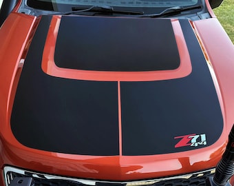 May include: An orange vehicle bonnet featuring a large matte black decal that covers the centre section. The decal includes a thin orange pinstripe down the middle and a red and white Z71 4x4 emblem positioned on the lower right side of the bonnet.