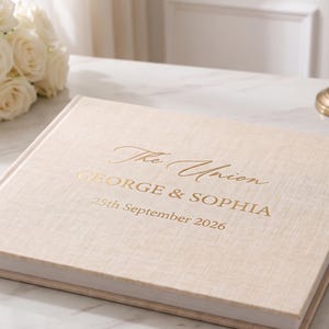 May include: A square, beige linen-covered photo album with gold foil lettering on the front cover that reads The Union, GEORGE & SOPHIA, and 25th September 2026. The book is closed and features a textured fabric finish on the exterior cover.