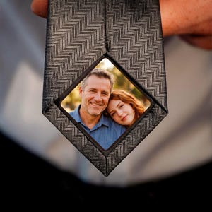 Peut inclure: Une cravate gris foncé avec un motif à chevrons subtil. La partie inférieure de la cravate présente un insert photographique carré en couleur montrant un homme d'âge mûr souriant et une jeune femme aux cheveux roux appuyée sur son épaule.