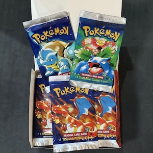 May include: A collection of four Pokémon Trading Card Game booster packs housed in an open cardboard box. The packs feature illustrations of Blastoise, Venusaur, and Charizard, with each pack labeled as containing 11 tradable game cards.