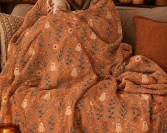 May include: A soft, burnt orange throw blanket featuring a repeating pattern of small white ghosts, delicate blue and green botanical sprigs, tiny stars, and small flowers. The fabric has a plush texture and covers the lap and shoulders.
