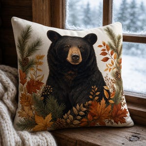 May include: A square throw pillow featuring a central illustration of a black bear surrounded by autumn foliage, including orange and brown maple leaves, pine needles, and small pinecones on a cream-colored fabric background.