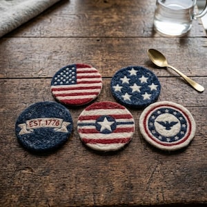 Op de afbeelding: Vijf ronde vilt onderzetters met patriottische ontwerpen in rood, wit en blauw. Eén toont de Amerikaanse vlag, een ander een sterrenveld, één heeft strepen met een centrale ster, één een adelaarsembleem en één bevat de tekst EST. 1776 op een lint.
