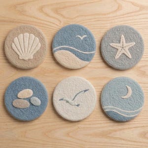 May include: A set of six circular felt coasters featuring coastal designs. The designs include a beige seashell, a blue and white wave with a bird, a blue starfish, three beige pebbles on blue, a white seagull, and a blue crescent moon with waves.