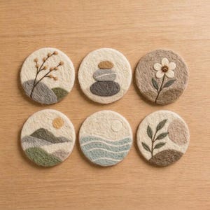 May include: A set of six circular felt coasters featuring embroidered designs including a branch, stacked stones, a flower, a mountain landscape with a sun, wavy water lines, and a leafy plant with a circular shape in neutral earth tones.
