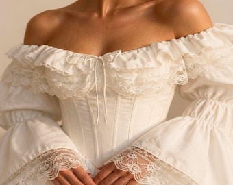 May include: An off-the-shoulder cream-colored blouse featuring a corset-style bodice with vertical seams, a ruffled neckline with a central drawstring tie, and long bell sleeves adorned with intricate white lace trim at the cuffs.