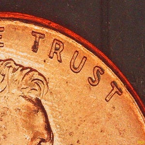 May include: A copper-colored coin featuring a portion of a human profile and the embossed text WE TRUST along the upper curved edge. The surface shows metallic texture and raised lettering against a dark background, capturing a close-up view.