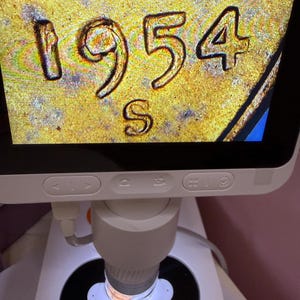 May include: A digital microscope displaying a magnified view of a gold-toned coin surface with the date 1954 and the mint mark S clearly visible. The device features a white base, a vertical stand, and a screen showing the metallic texture.