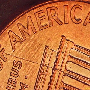 May include: A macro view of a copper-coloured United States one-cent coin showing the text OF AMERICA and a portion of the Lincoln Memorial building. A distinct crack runs diagonally across the surface of the metal coin near the lettering.