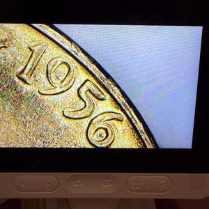 May include: A digital microscope screen displaying a magnified view of a metallic coin surface. The gold-toned metal shows the embossed year 1956 with visible texture and raised numerals. The screen is housed in a white plastic frame with control buttons.