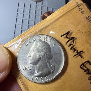 Puede incluir: Una moneda de cuarto de dólar Washington de 1966 dentro de una funda de plástico transparente unida a un soporte de papel color canela. La moneda muestra el perfil de George Washington, la palabra LIBERTY, la frase IN GOD WE TRUST y el año 1966.
