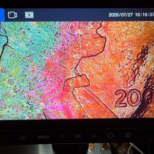 Pode incluir: Uma tela de microscópio digital exibindo uma superfície texturizada altamente ampliada com tons vibrantes de verde, laranja e rosa. A imagem mostra o ano 2001 e a letra R impressos na superfície, juntamente com várias rachaduras e irregularidades.