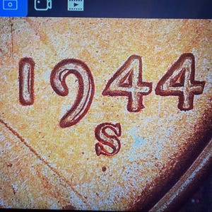 Könnte beinhalten: Eine Nahaufnahme der kupferfarbenen Münzoberfläche mit dem geprägten Datum 1944 und dem Münzzeichen S direkt unter der Jahreszahl. Die metallische Textur zeigt feine Oberflächendetails und erhabene Schriftzeichen in einem warmen Bronzeton.