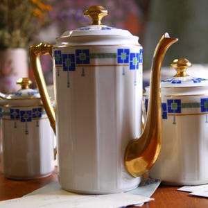 Op de afbeelding: Een witte porseleinen koffiepot en twee bijpassende potten met deksel, voorzien van verticale groeven, goudkleurige handvatten en knoppen, en een geometrisch patroon van blauwe en groene vierkanten met dunne gouden lijnen langs de rand.