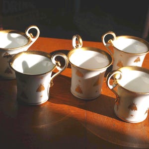 May include: A set of four white porcelain cups featuring gold-colored moth motifs and gold-trimmed rims. Each cup has a curved, gold-accented handle and a smooth, flared body. The exterior surface is decorated with small, repeating insect patterns.