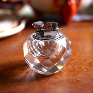 Peut inclure: Un briquet de table en verre transparent à facettes avec un corps sphérique taillé en diamant. Le mécanisme supérieur est en métal argenté poli, doté d'un bouton d'allumage circulaire et d'un petit levier latéral pour le réglage.
