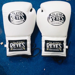 Peut inclure: Une paire de gants de boxe blancs avec des lacets noirs. Chaque gant présente un logo noir circulaire sur le poignet et le dos de la main avec le texte Cleto Reyes Hecho en Mexico et le mot Profesional sur l'étiquette du poignet.