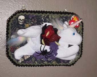 Op de afbeelding: Een witte pluche eenhoorn gemonteerd op een rechthoekige plaat met een donkere bloemenachtergrond. De eenhoorn heeft manen en een staart in regenboogkleuren, een rood hartvormig ornament, een zwarte spin en een rood Ty-label.